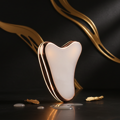 Luxury electronic Gua Sha beauty instrument with soft glow on dark moody background with gold accent lighting